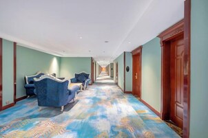 A hotel hallway with a patterned carpet, blue upholstered seating, and wooden doors.