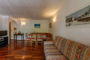 A living room with a wooden dining table, chairs, a sofa, a television, and a painting on the wall.