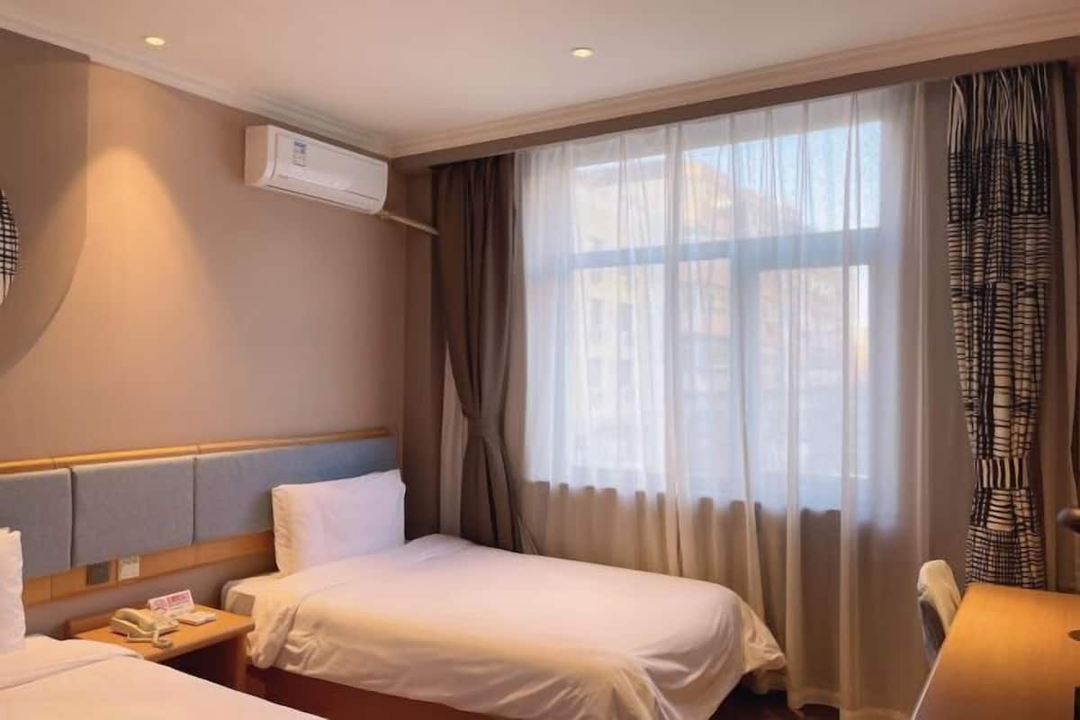 Une chambre d’hôtel avec deux lits, un bureau, une chaise, un appareil de climatisation et une fenêtre avec des rideaux.