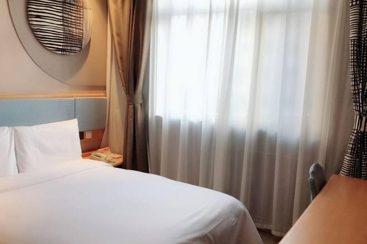 Une chambre d’hôtel avec un lit, une table de chevet, un téléphone et une fenêtre avec des rideaux légers.