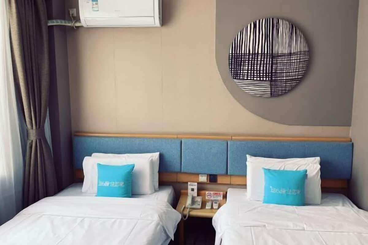 Une chambre d’hôtel avec deux lits, chacun étant équipé de draps blancs et d’un oreiller bleu. Un climatiseur est fixé au mur, et une décoration circulaire orne le mur au-dessus des lits.