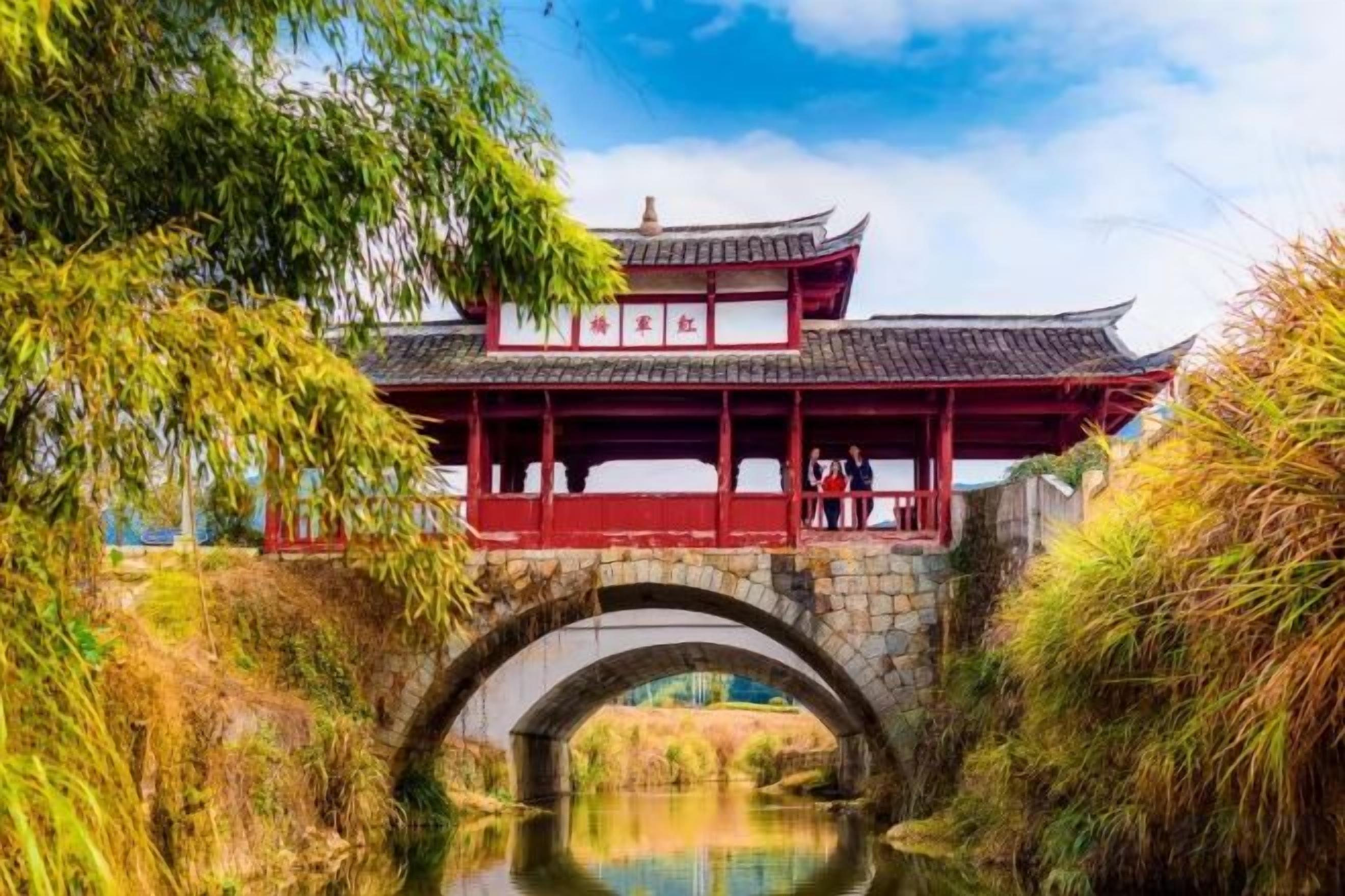 Ein traditioneller chinesischer Pavillon über einer Stegbrücke, im Vordergrund ein ruhiges Gewässer.