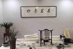 Una habitación con una obra de caligrafía enmarcada en la pared, una planta en maceta, una silla y una mesa con ropa de cama doblada y varios objetos.