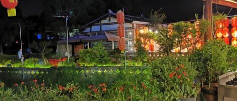 A nighttime view of a traditional building with a garden area featuring potted plants and a pond.