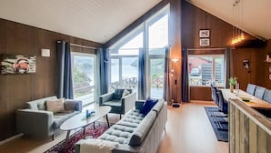 Un salon spacieux avec un grand canapé, une table basse et un coin repas comprenant une table et des chaises. De grandes fenêtres offrent une vue sur l’extérieur.