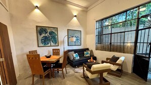 Un salon chaleureux avec un coin repas, comprenant une table et des chaises en bois, un canapé et une grande fenêtre à barreaux.