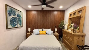 Une chambre à coucher comprenant un lit, une tête de lit en bois, des tables de chevet, une commode, un ventilateur de plafond et un tableau encadré accroché au mur.