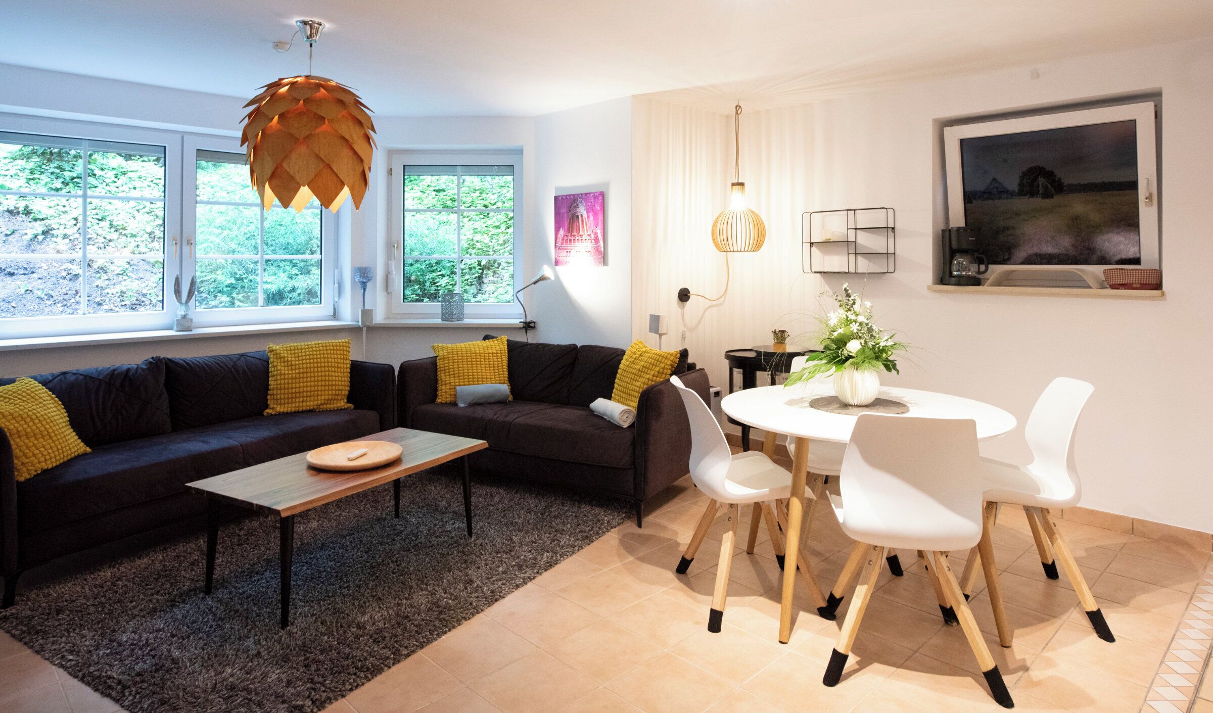 A modern living room with a sofa, coffee table, and dining area with chairs. There are large windows with a view of greenery outside.