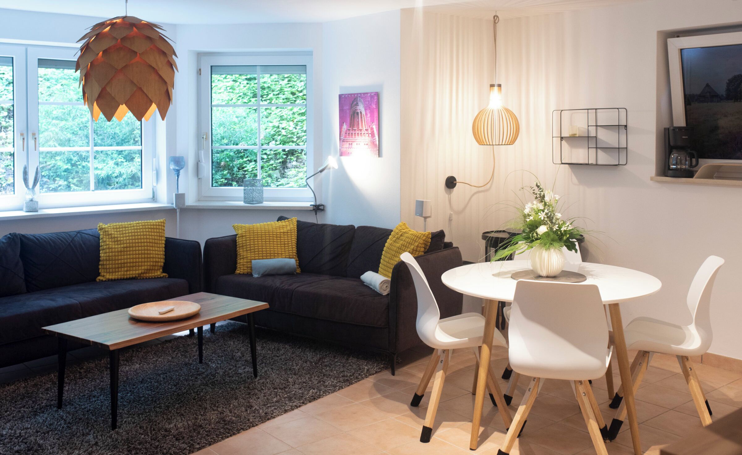 Ett modernt vardagsrum med en soffa, ett soffbord och ett matbord. Det finns fönster med gardiner, en hängande lampa och en inramad tavla på väggen.