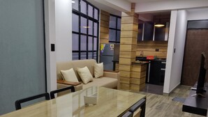 A modern living room with a glass-top dining table, a beige sofa, and a kitchen area with wooden accents.