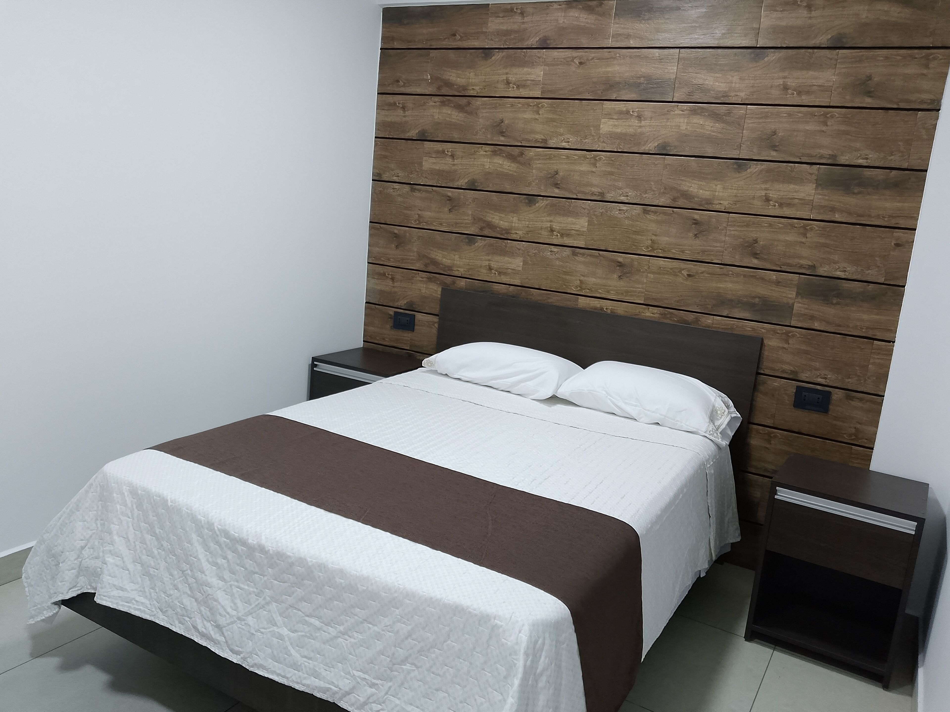 A neatly made bed with a white and brown bedspread, flanked by two bedside tables with dark wood finishes.