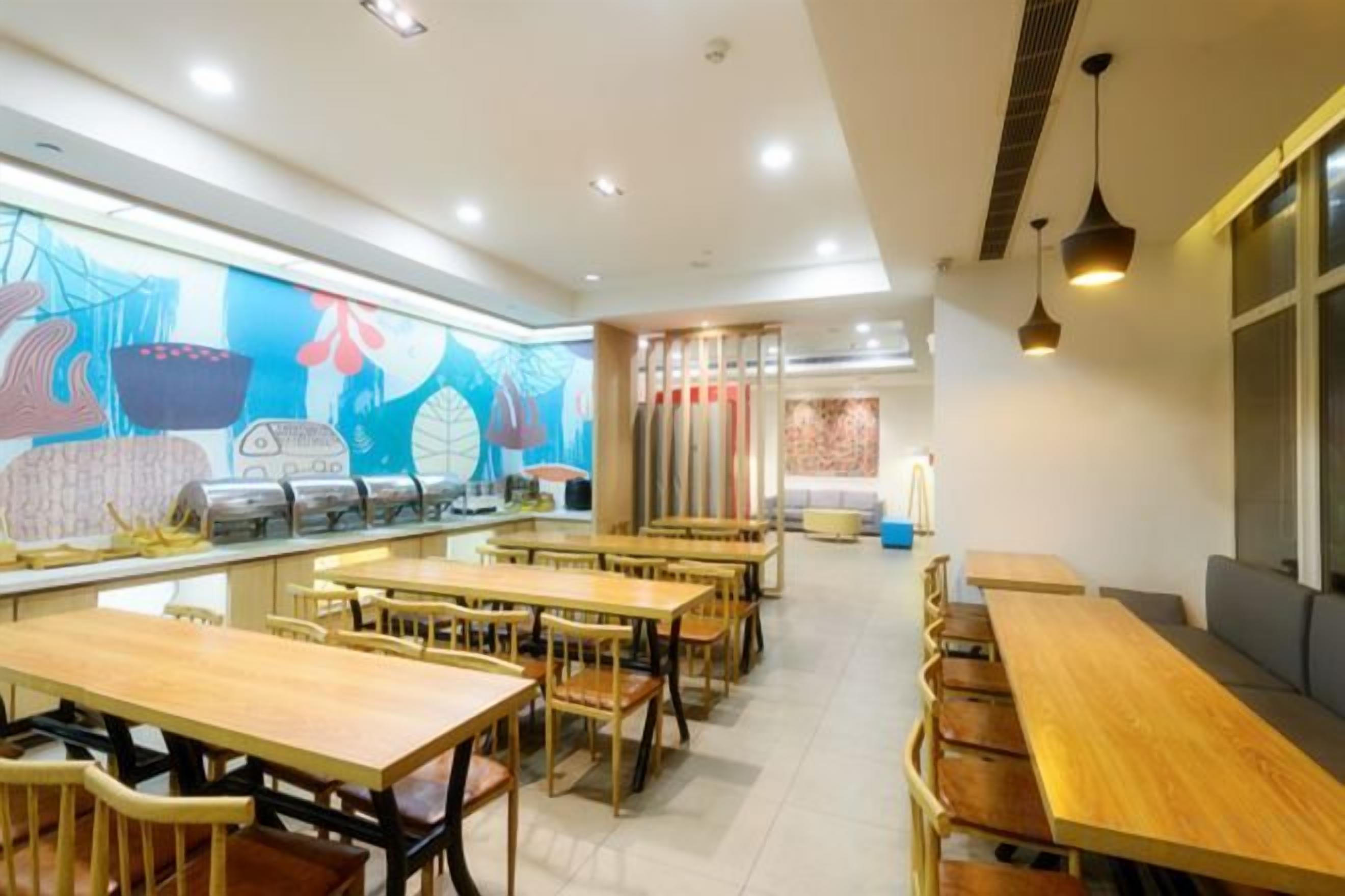 A restaurant with multiple wooden tables and chairs, a colorful mural on the wall, and pendant lighting.