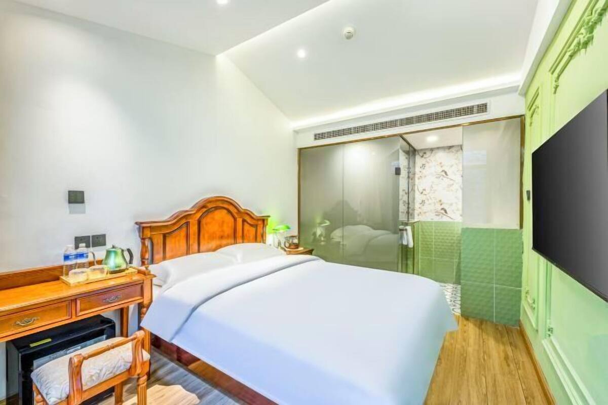 Une chambre d’hôtel avec un lit en bois, un bureau avec une chaise, une télévision et une salle de bain dotée d’une douche à parois de verre.