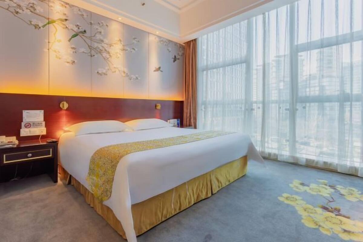 A hotel room with a large bed, a nightstand, a wall-mounted telephone, and a window with sheer curtains.
