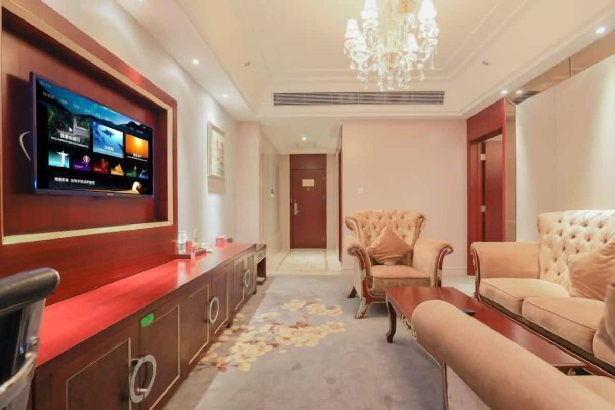A hotel lobby with a flat-screen TV mounted on the wall, a reception desk, a sofa, a chair, and a chandelier.