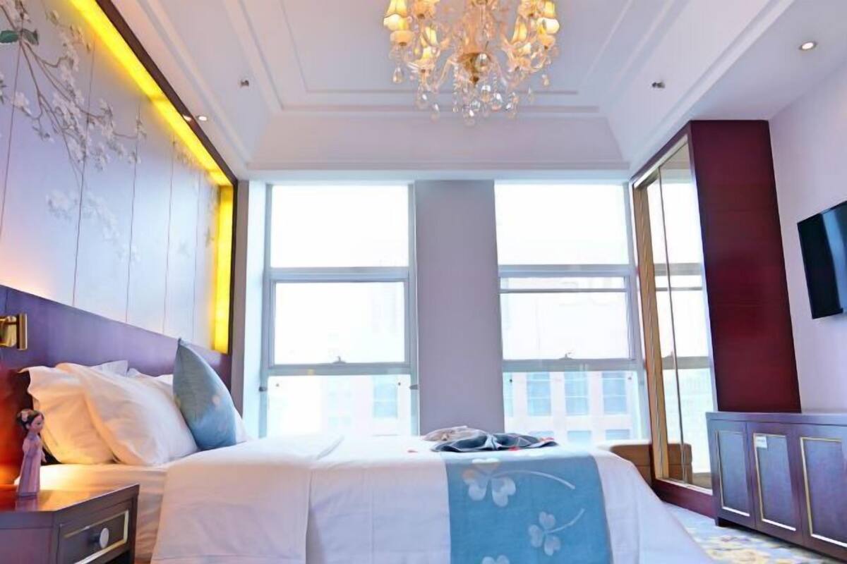 A hotel room with a large bed, a chandelier, a dresser, and a TV on the wall.