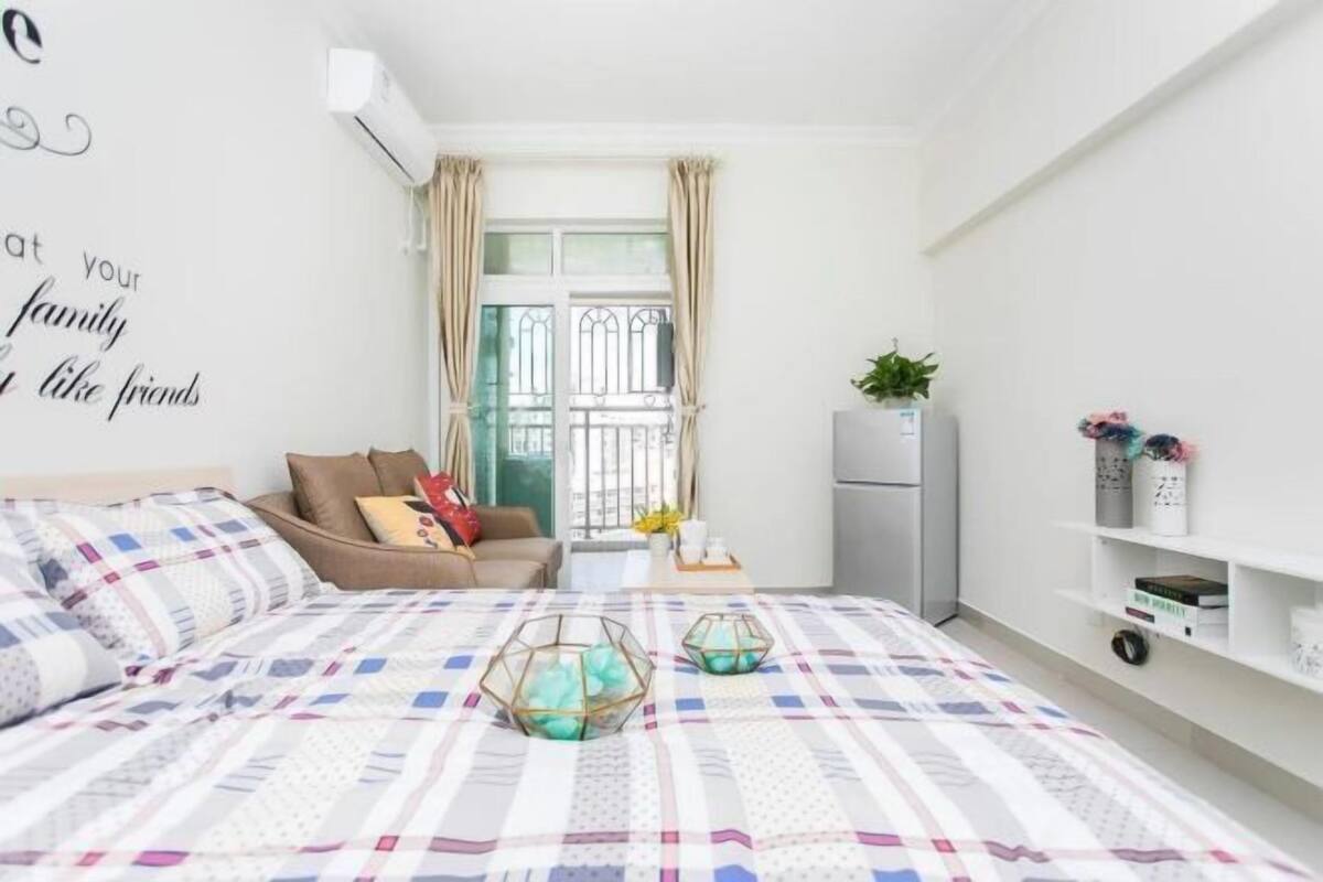 Une chambre à coucher avec un lit, un canapé, une fenêtre avec des rideaux, une plante et un mur orné d’une citation.