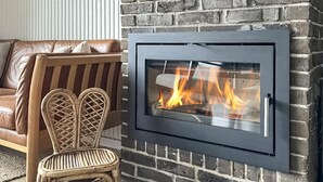 Una chimenea moderna con frente de vidrio, ubicada frente a una pared de ladrillo, junto a un sofá de cuero marrón y una silla de mimbre.