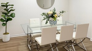 Ein moderner Essbereich mit einem Glastisch, weißen Stühlen, einer Topfpflanze, einer Vase mit Blumen und einem runden Spiegel an der Wand.