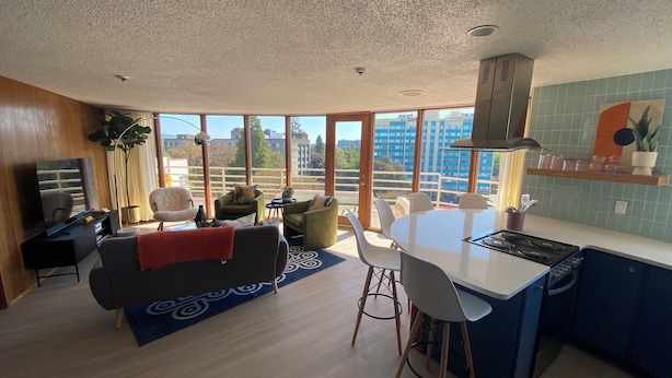 A modern living room with a sofa, chairs, a dining table, and a kitchen area with a stove. Large windows offer a view of buildings outside. - Newly renovated penthouse getaway! (Eugene)