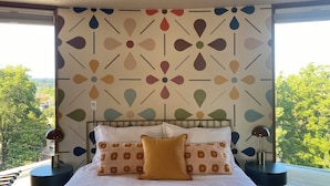 A bedroom with a patterned wall, a bed with white and patterned pillows, and a view of trees and buildings through a window. - Newly renovated penthouse getaway! (Eugene)