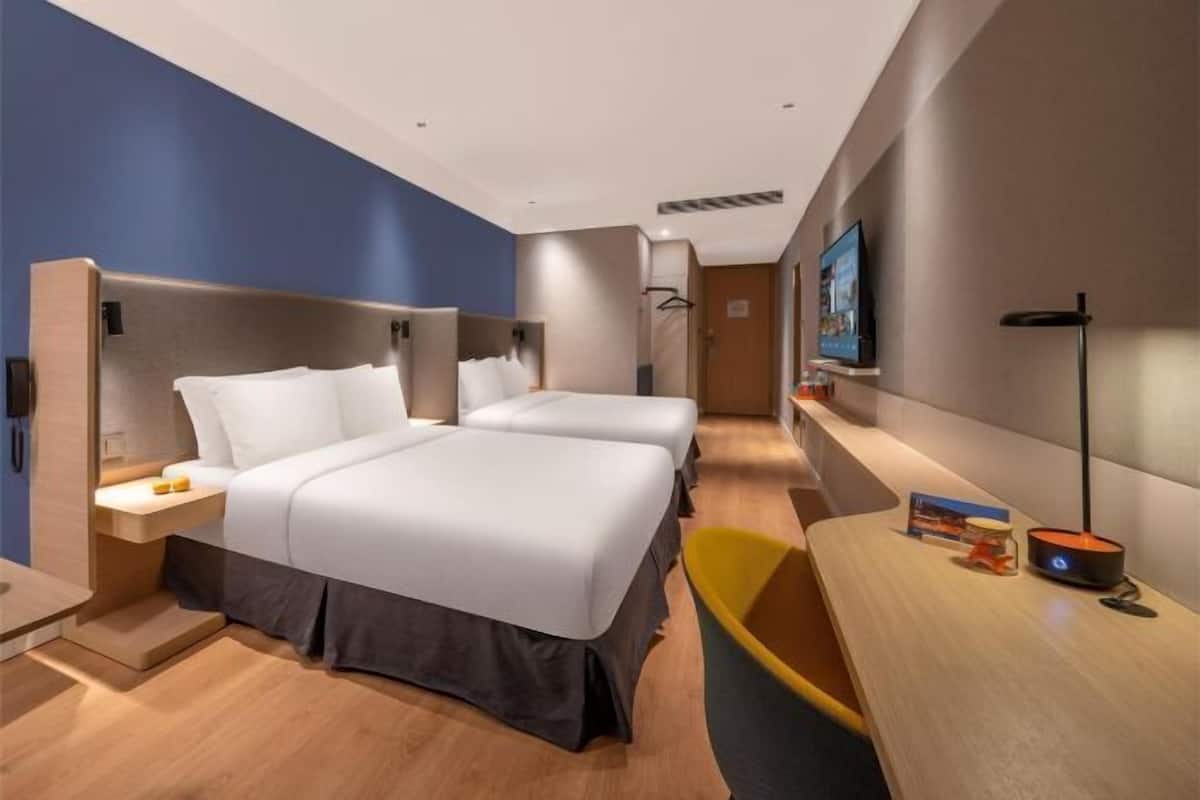 A hotel room with two beds, a desk, a chair, a TV, and a bedside table with a lamp.