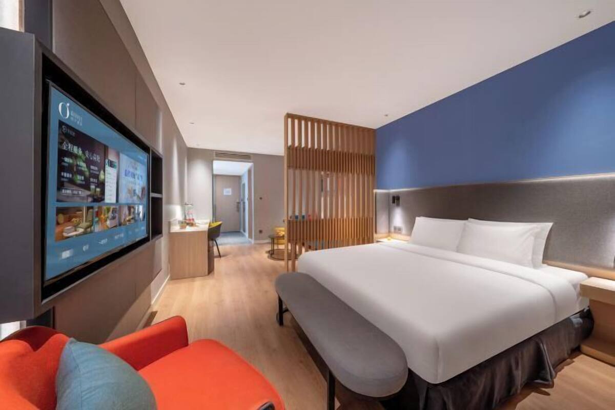 A modern hotel room with a large bed, a TV mounted on the wall, a desk, and a seating area with a bench.
