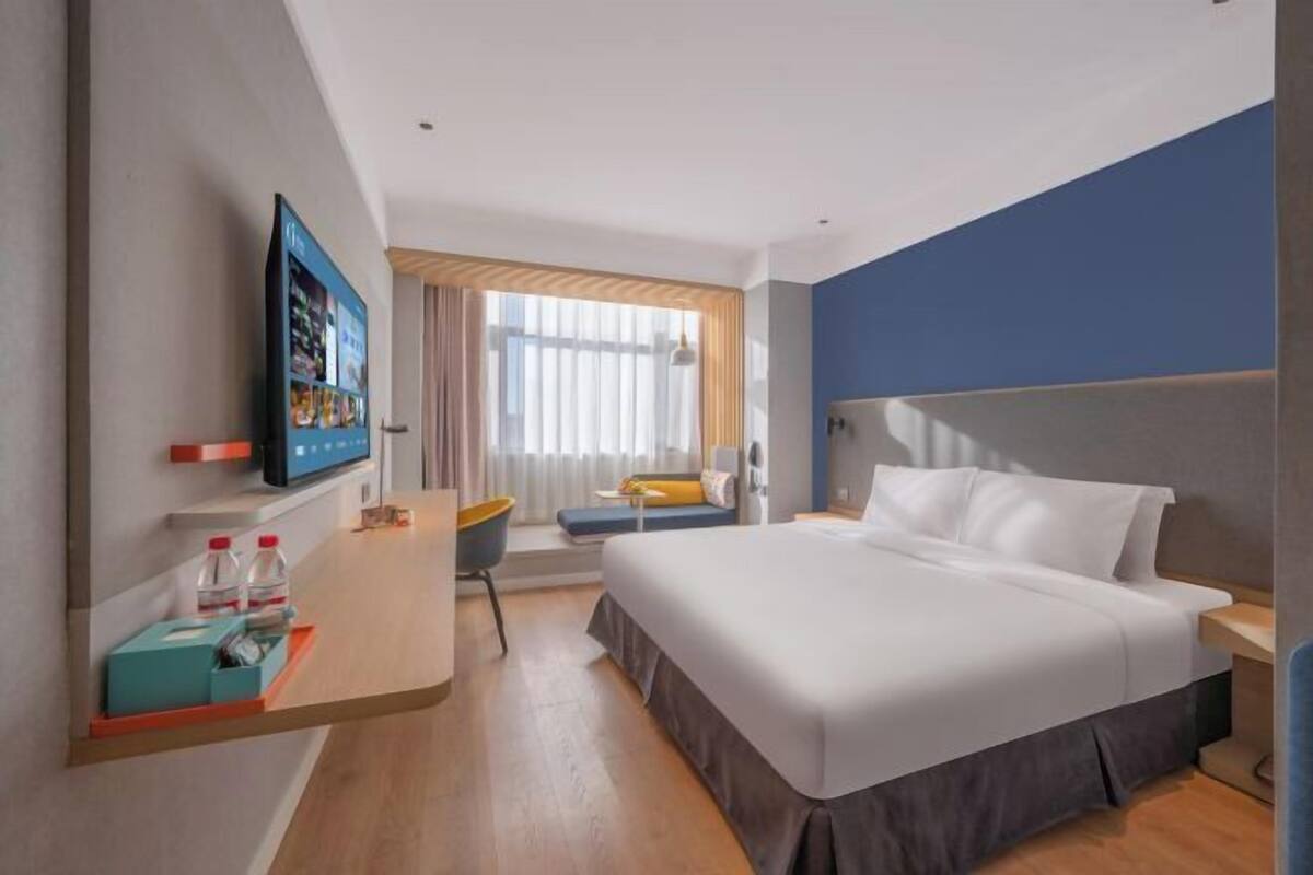 A hotel room with a bed, a TV, a desk with a chair, a window with curtains, and a blue accent wall.