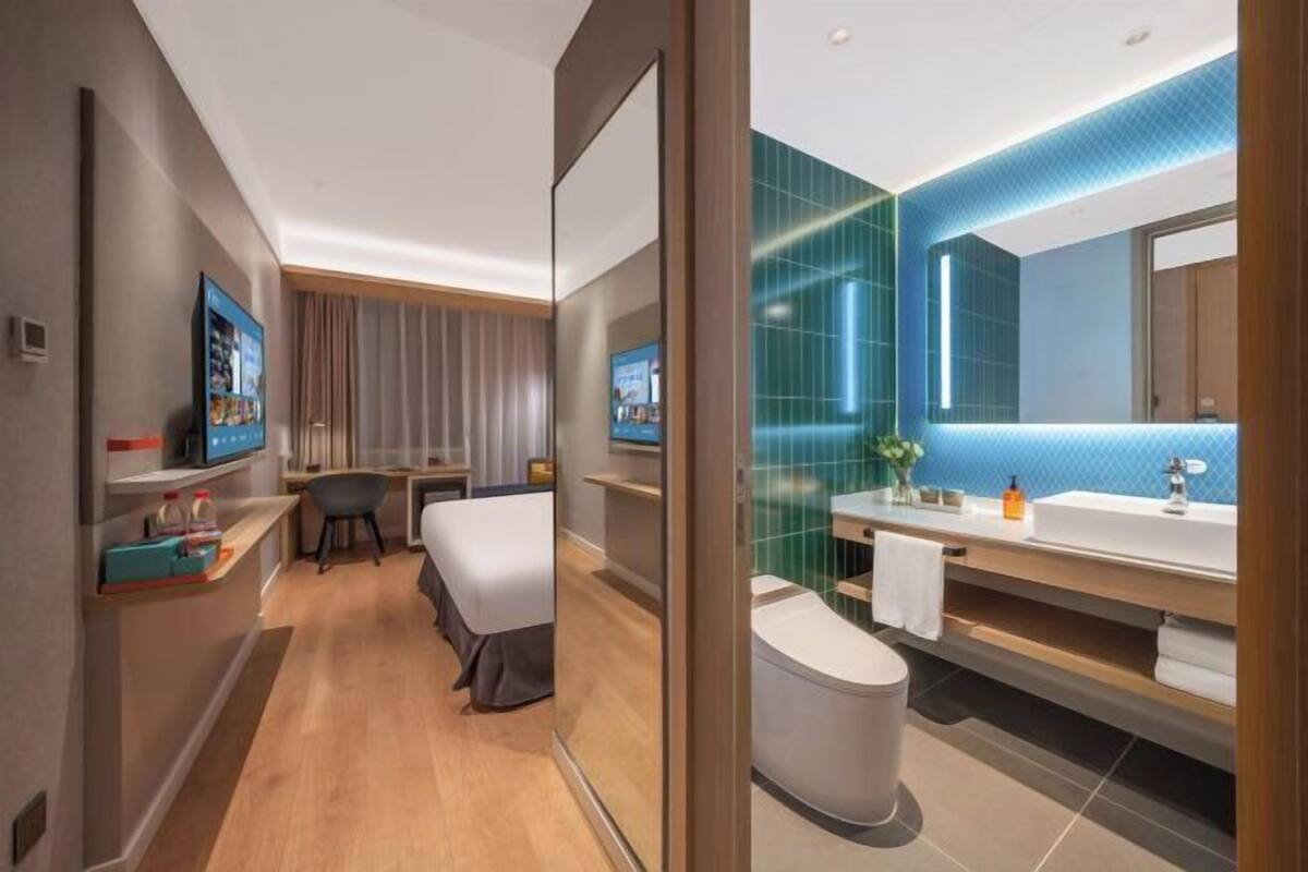 A modern hotel room with a bed, a desk, a television, a bathroom with a large mirror, and a shower.