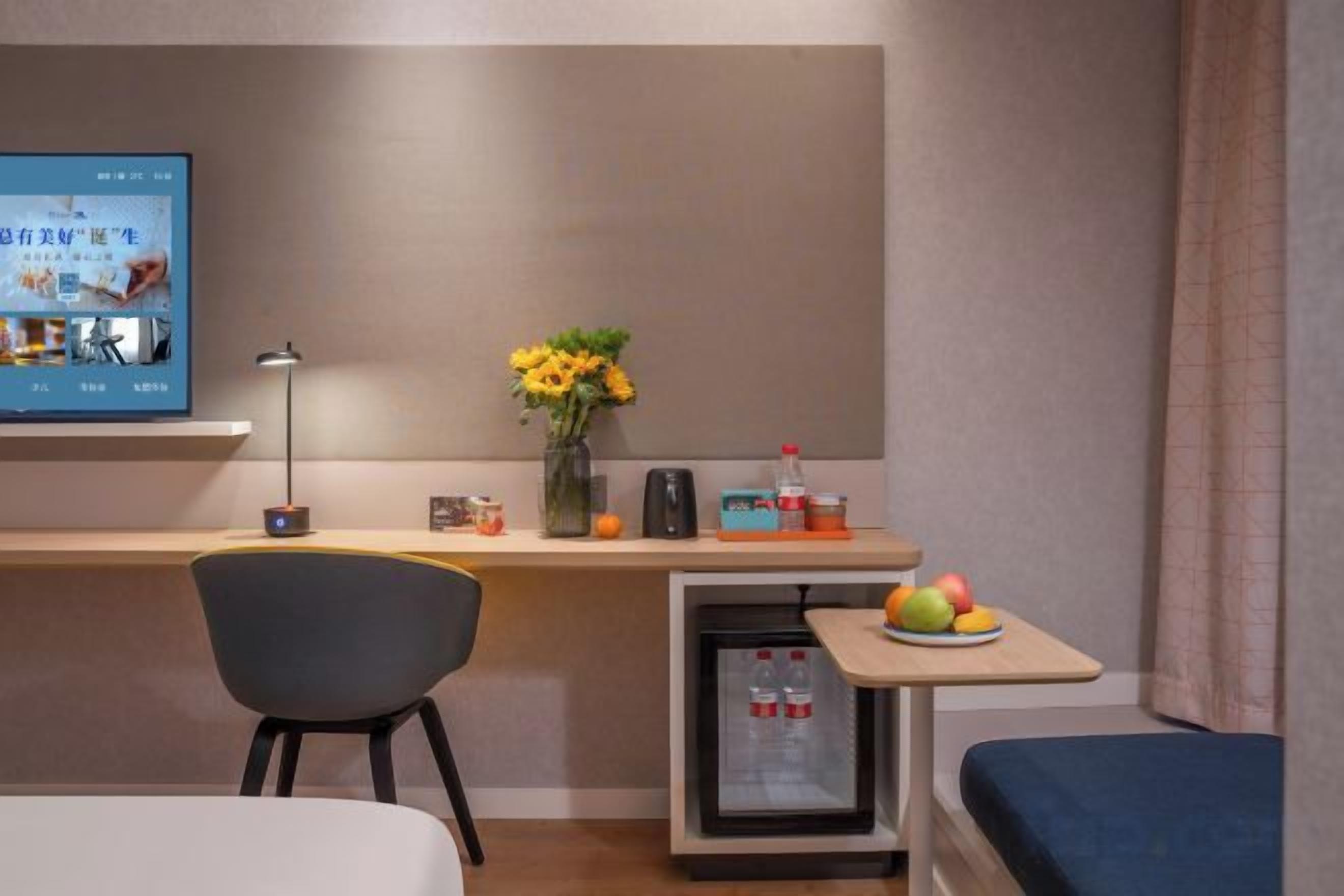 A hotel room with a bed, a desk with a computer, a chair, a small table with fruit, and a refrigerator.