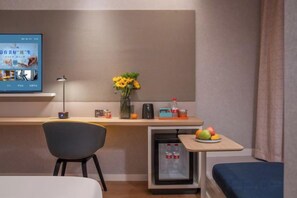 Une chambre d’hôtel comprenant un lit, un bureau avec un ordinateur, une chaise, une petite table avec des fruits et un réfrigérateur.