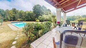 Un patio cubierto con piscina, mobiliario exterior y vistas a un área boscosa.