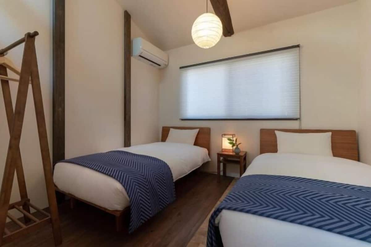 A room with two beds, a wooden ladder, a small table with a lamp, and a window with blinds.