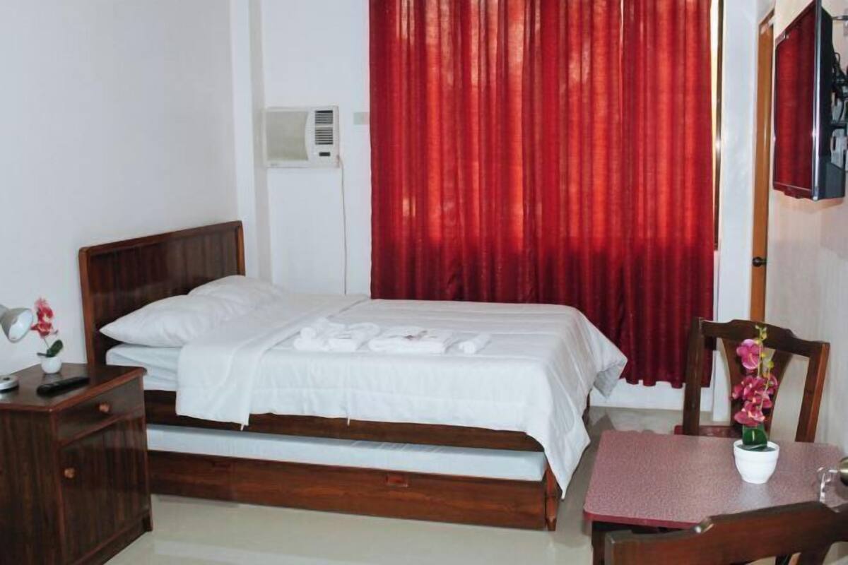 Una cama individual con cabecera de madera, una mesita de noche con lámpara y control remoto, una pequeña mesa con un florero y una cortina roja.