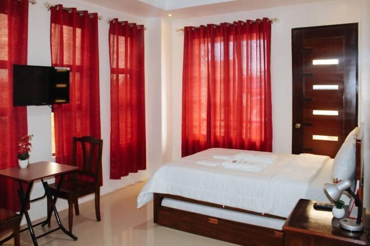 Un dormitorio con cama, escritorio, silla, televisor y cortinas rojas.
