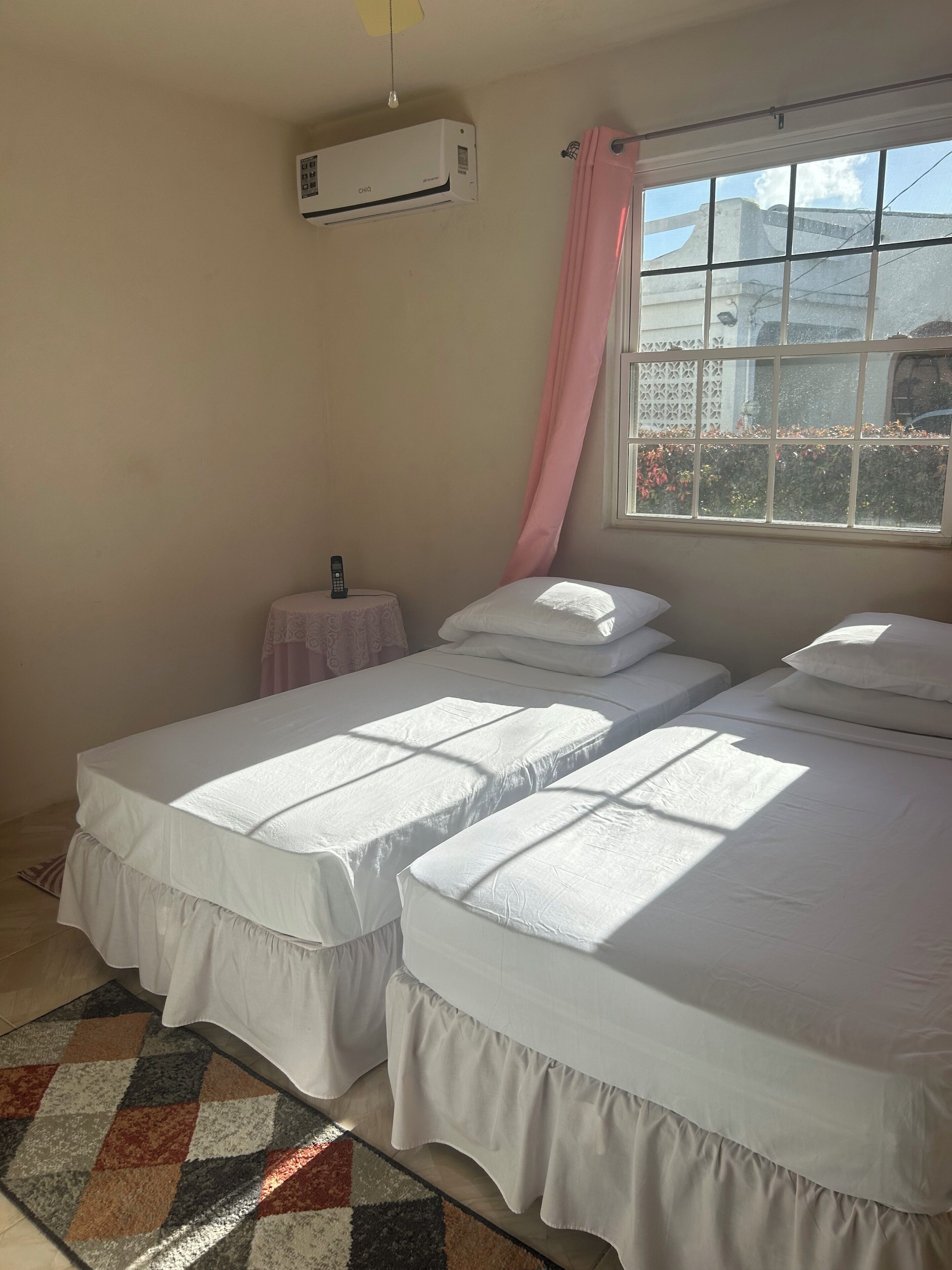 A room with two beds, a small table, an air conditioner, and a window with a view of the outside.