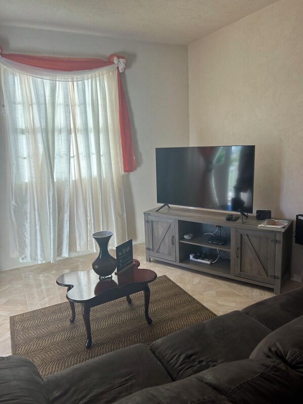 A living room with a flat-screen TV on a wooden cabinet, a dark-colored sofa, a small table with a vase, and sheer curtains with a red accent. - StayHaven Escapes (Saint Thomas)
