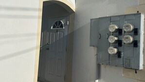 Een gebouw met een witte gevel, een grijze technische kast met meters en een witte deur met een gebogen bovenkant.