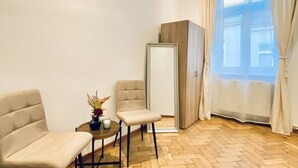 Una habitación con dos sillas color beige, una mesita redonda con un florero, un mueble de madera y vistas a través de cortinas translúcidas.