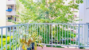 Un balcón con baranda metálica, una planta en maceta y vista a un edificio de varios pisos con balcones.