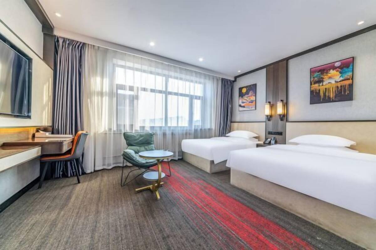 Hotelkamer met twee bedden, een bureau, een stoel, een klein rond tafeltje en een groot raam met gordijnen.