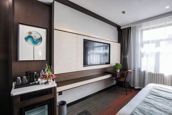 A modern hotel room with a large bed, a TV mounted on the wall, a desk with a chair, and a small table with a plant.