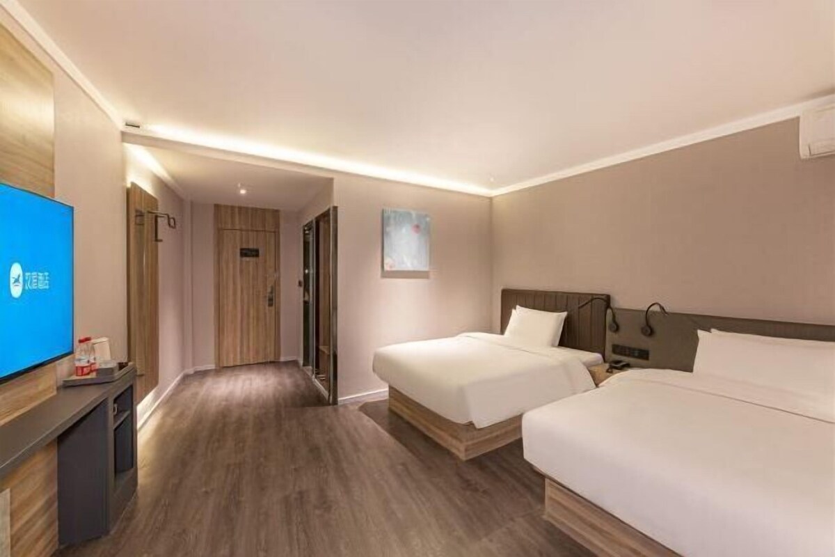 A hotel room with a wooden floor, two beds, a flat-screen TV, and a hallway leading to another room.