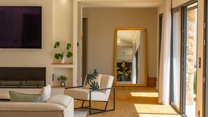 Una sala de estar moderna con un sofá, un sillón y un espejo grande. Hay un televisor montado en la pared y una planta en una estantería.