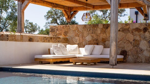 Un área junto a la piscina con una pérgola de madera, pared de piedra y un sofá blanco con cojines.