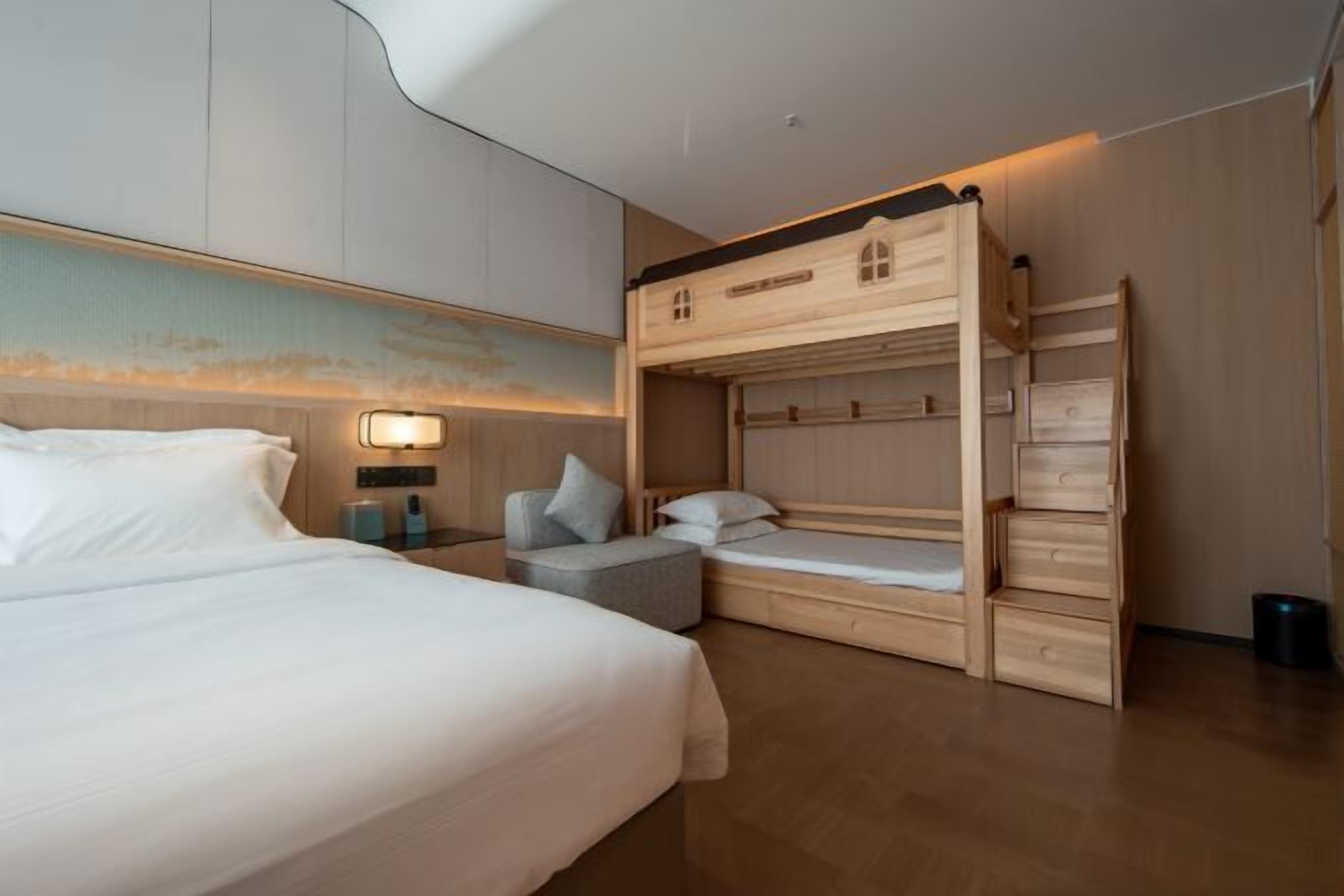 Moderni hotellihuone, jossa on suuri sänky, kerrossänky, pieni oleskelualue ja seinään kiinnitetty valaisin.