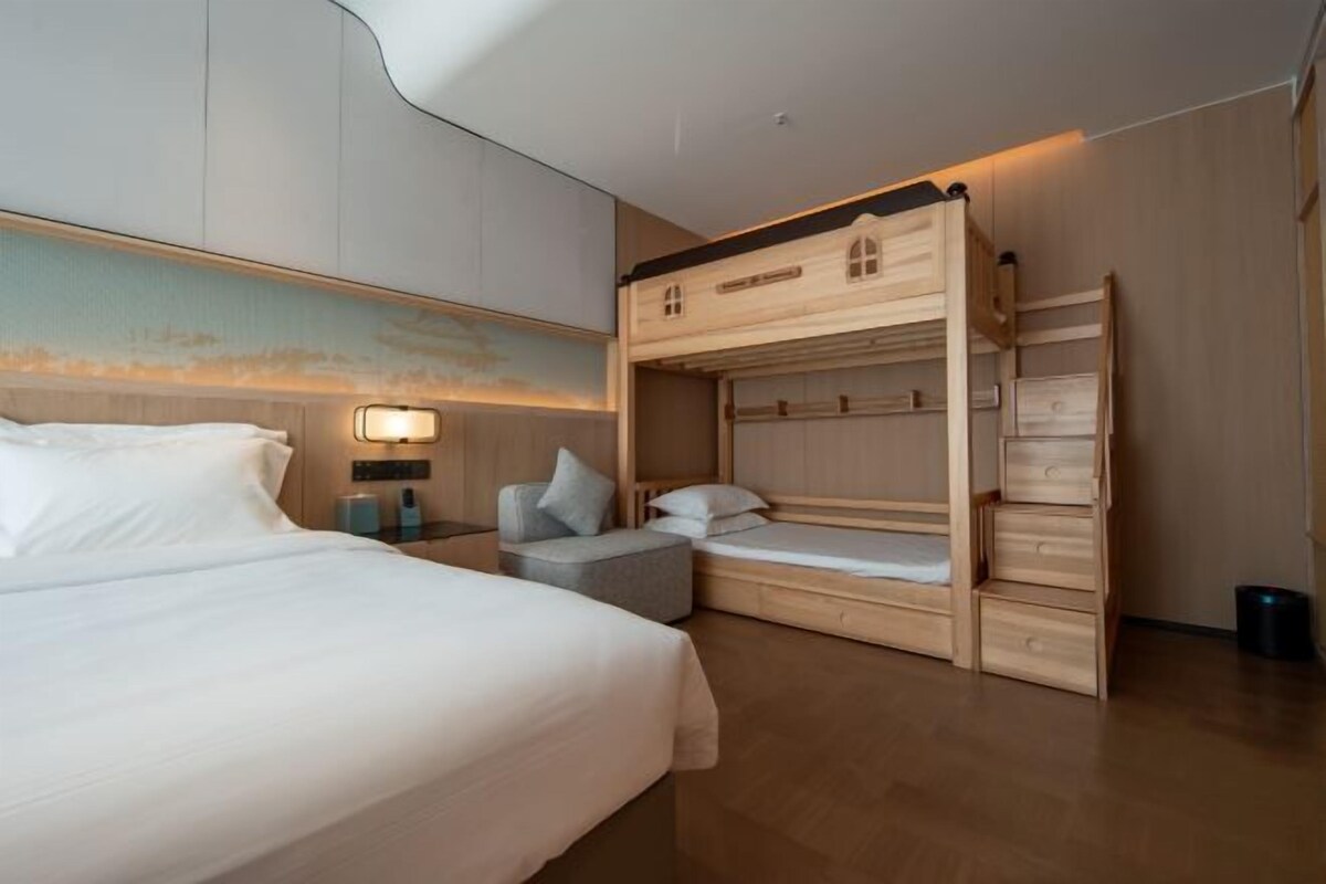 Una habitación de hotel moderna con una cama grande, una litera, un pequeño rincón de descanso y una lámpara fijada en la pared.