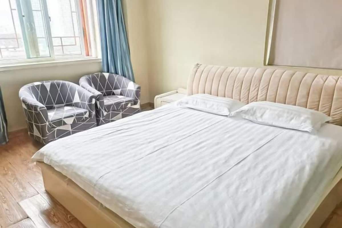 Une chambre à coucher avec un lit, deux fauteuils, une fenêtre avec des rideaux et un téléviseur fixé au mur.