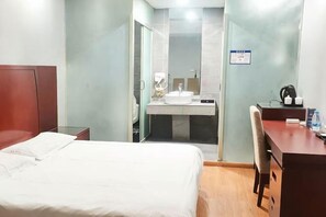 Une chambre d’hôtel comprenant un lit, un bureau, une chaise, une salle de bain avec un lavabo et un miroir, et une douche.