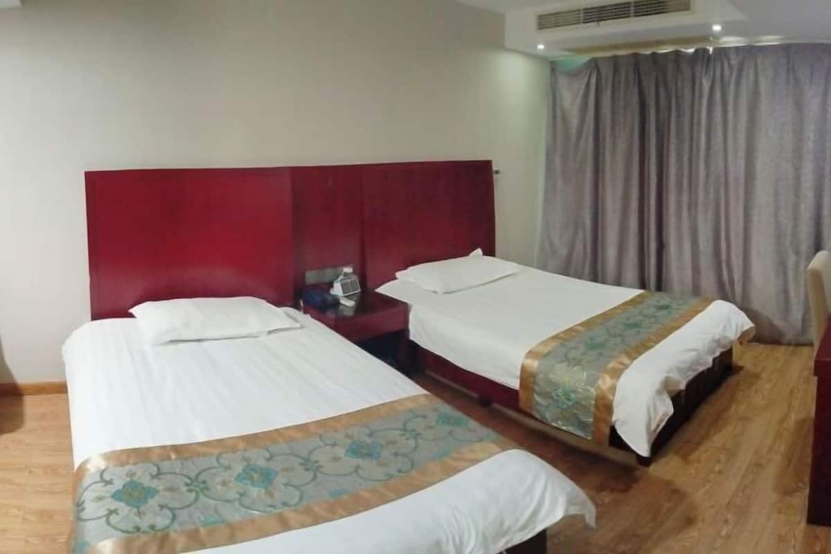 Une chambre d’hôtel avec deux lits, une tête de lit rouge, une table de chevet avec une lampe et une fenêtre avec des rideaux.
